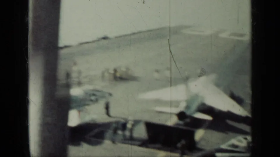 1962 Cuba home-movie frame showing a carrier deck with parked jets, crew figures, railing, open water, and a faint distant shoreline.