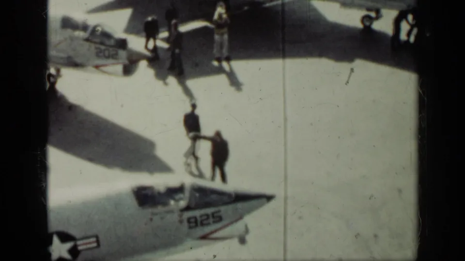 High-angle Cuba 1962 frame showing personnel on a bright tarmac beside U.S. Navy fighter jets with visible numbers 202 and 925.
