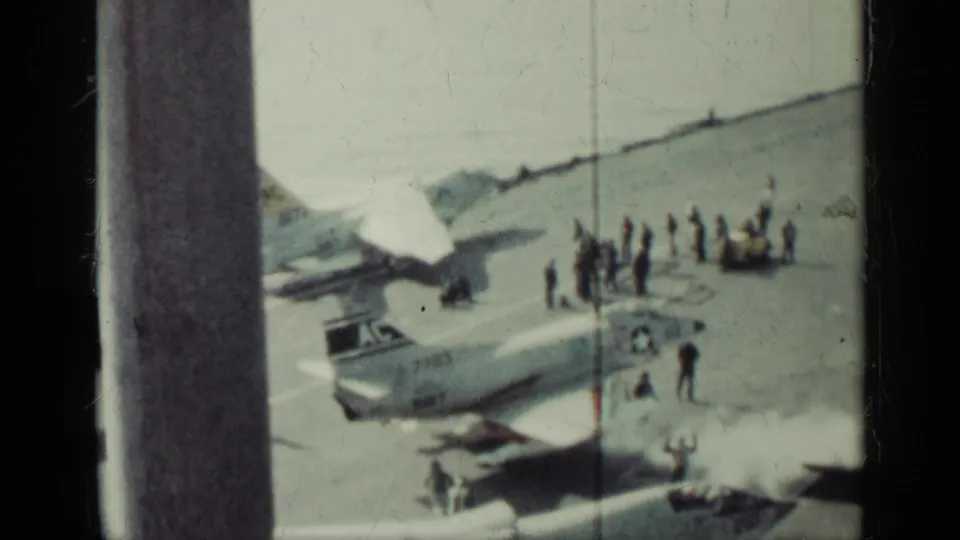 Home-movie view of a carrier flight deck with U.S. Navy jets, crew figures, and light smoke or exhaust around the aircraft.