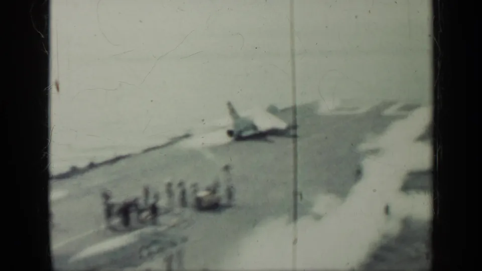 A soft 8mm frame shows a jet on an aircraft carrier deck, with crew grouped nearby and smoke or exhaust drifting across the scene.