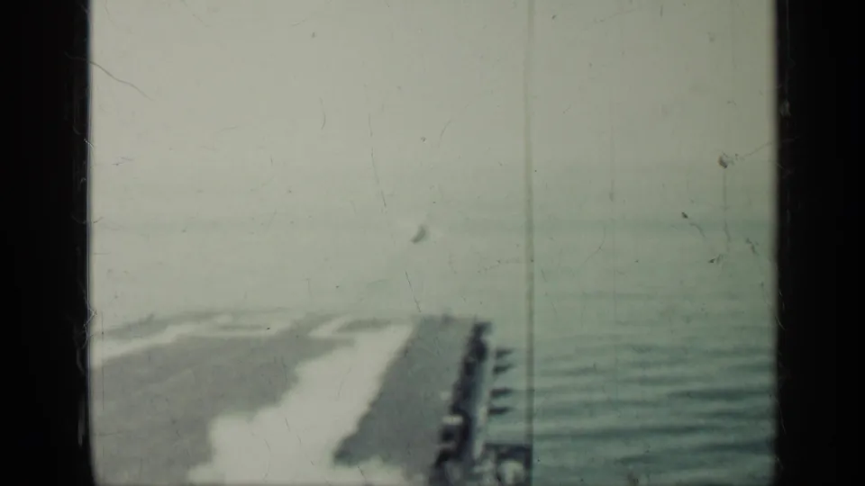 Final 8mm frame looking down a carrier deck toward open water, with a small aircraft shape ahead and deck edge visible at sea.