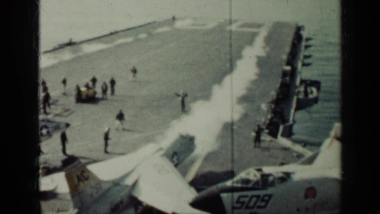 Jet aircraft and deck crew on a carrier flight deck beside the sea.