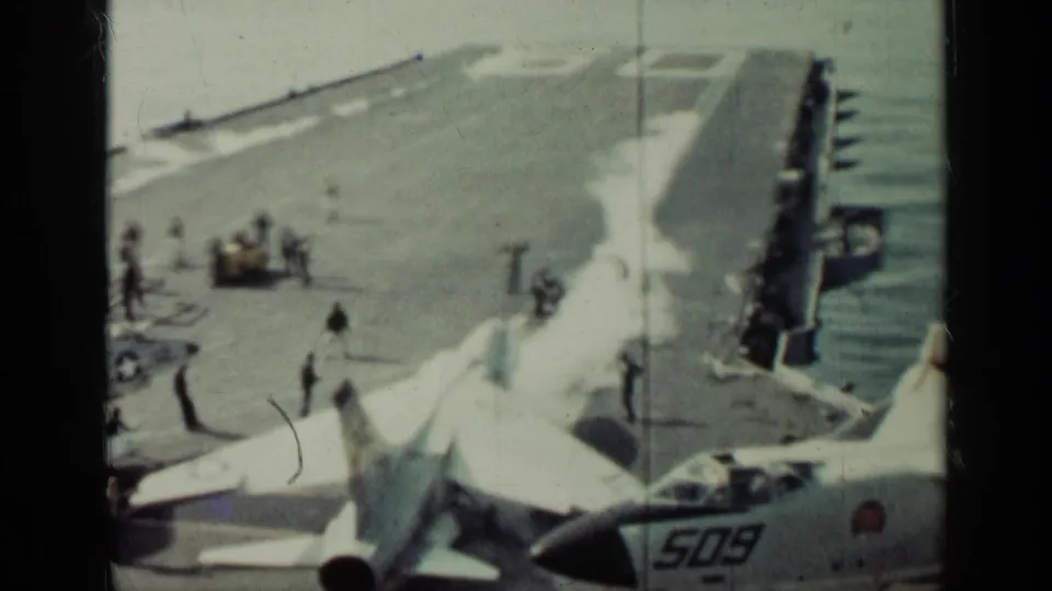 Grainy 1962 Cuba-related frame of U.S. Navy carrier deck activity, with jets in the foreground and crew scattered across the deck.