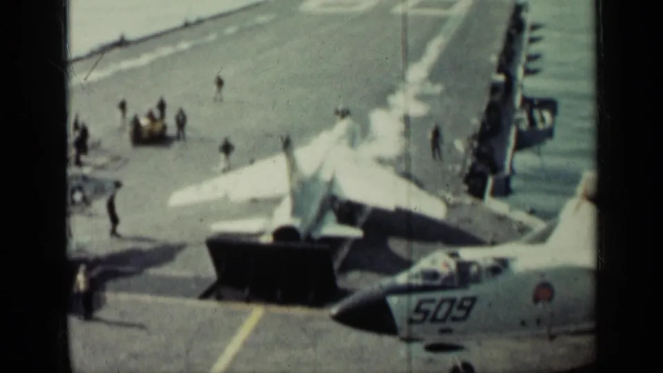 Carrier-deck jet preparation with a raised blast deflector, nearby crew, and a marked aircraft nose visible at the lower right.