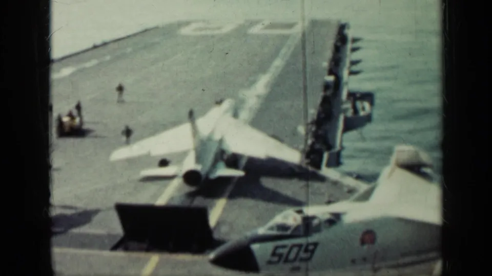 Wide 8mm view of a carrier deck at sea, showing a jet aligned near a raised deflector with crew figures and aircraft markings visible.
