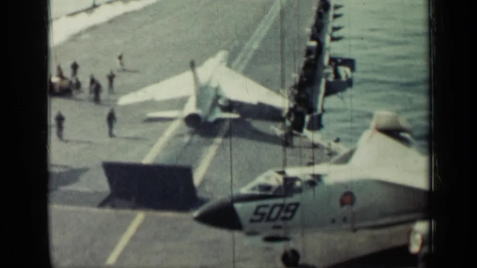 Carrier flight deck scene with a jet nose number, crew figures, ocean horizon, and launch-area markings in 1962-supported footage.