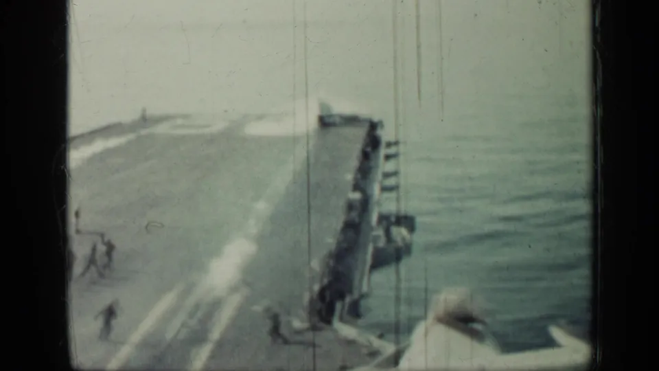Soft 8mm frame showing a jet departing or moving along the carrier deck, with crew figures and ocean alongside the ship.