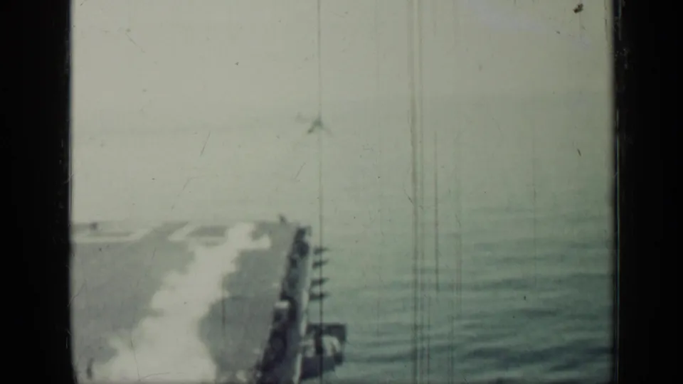 Faded 8mm view over the carrier deck and ocean, with a small aircraft shape in the distance and deck edge visible.