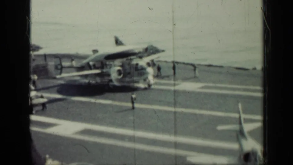 A military airplane sits on a carrier flight deck near the ocean, with small deck crew figures visible in the 1962 Cuba-linked footage.