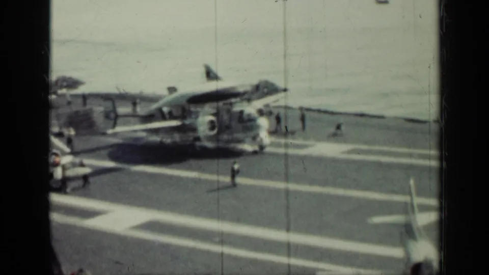 1962 Cuba-linked 8mm frame showing a military aircraft on a carrier deck, painted flight lines, ocean water, and small crew figures.