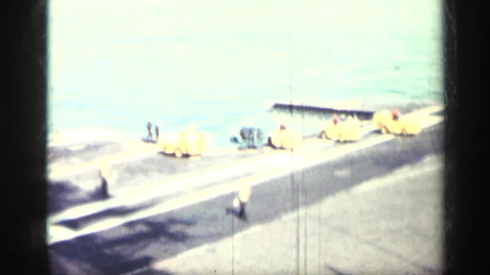 Distant uniformed men work near yellow deck equipment on an aircraft-carrier deck by the sea, matching the supplied Cuba 1962 metadata.