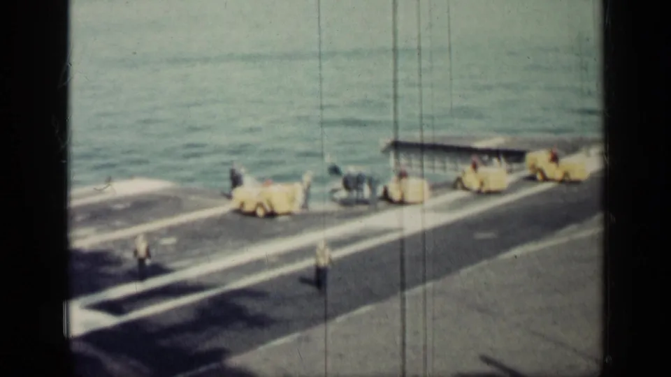 Wide 8mm view of men on a carrier deck near yellow equipment and a small covered station, with open sea behind them.