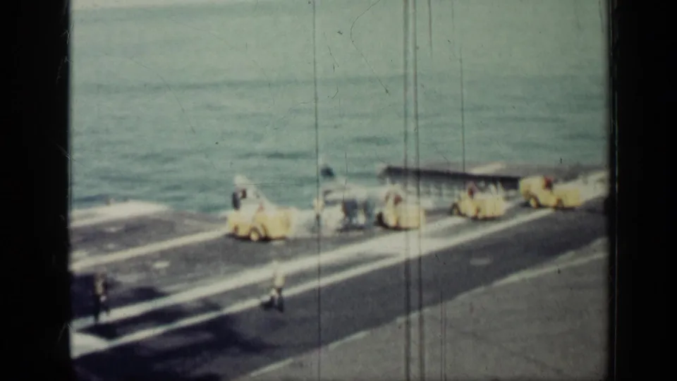 Uniformed men appear to work around yellow deck equipment on an aircraft-carrier deck, with sea and faded 8mm film wear visible.