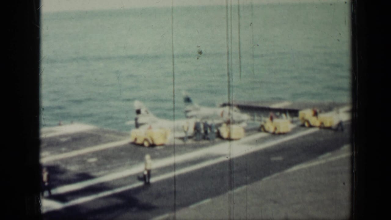 Jet and deck crew on an aircraft carrier deck with yellow support vehicles near the ocean.