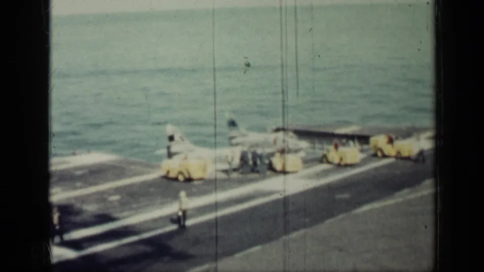 1962 Cuba-era 8mm frame showing a jet on an aircraft carrier deck as crew and yellow support vehicles work near the sea.