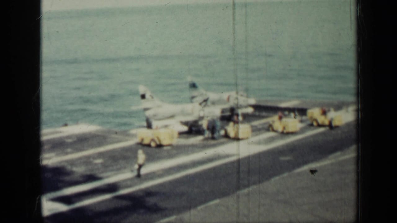 Carrier deck scene with jet, crew members, yellow vehicles, and open water behind the ship.