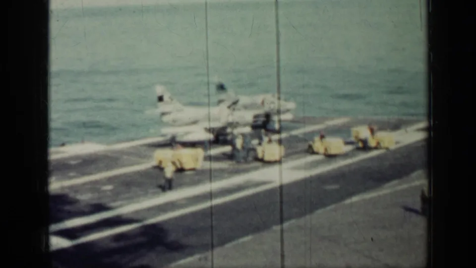 1962 Cuba-era 8mm view of a carrier flight deck as sailors guide activity around a jet and nearby yellow vehicles.