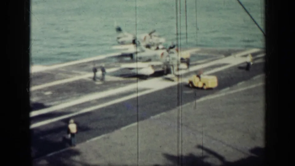 A 1962 Cuba-era home-movie frame shows carrier deck crew near a jet, with a yellow support vehicle and sea behind them.