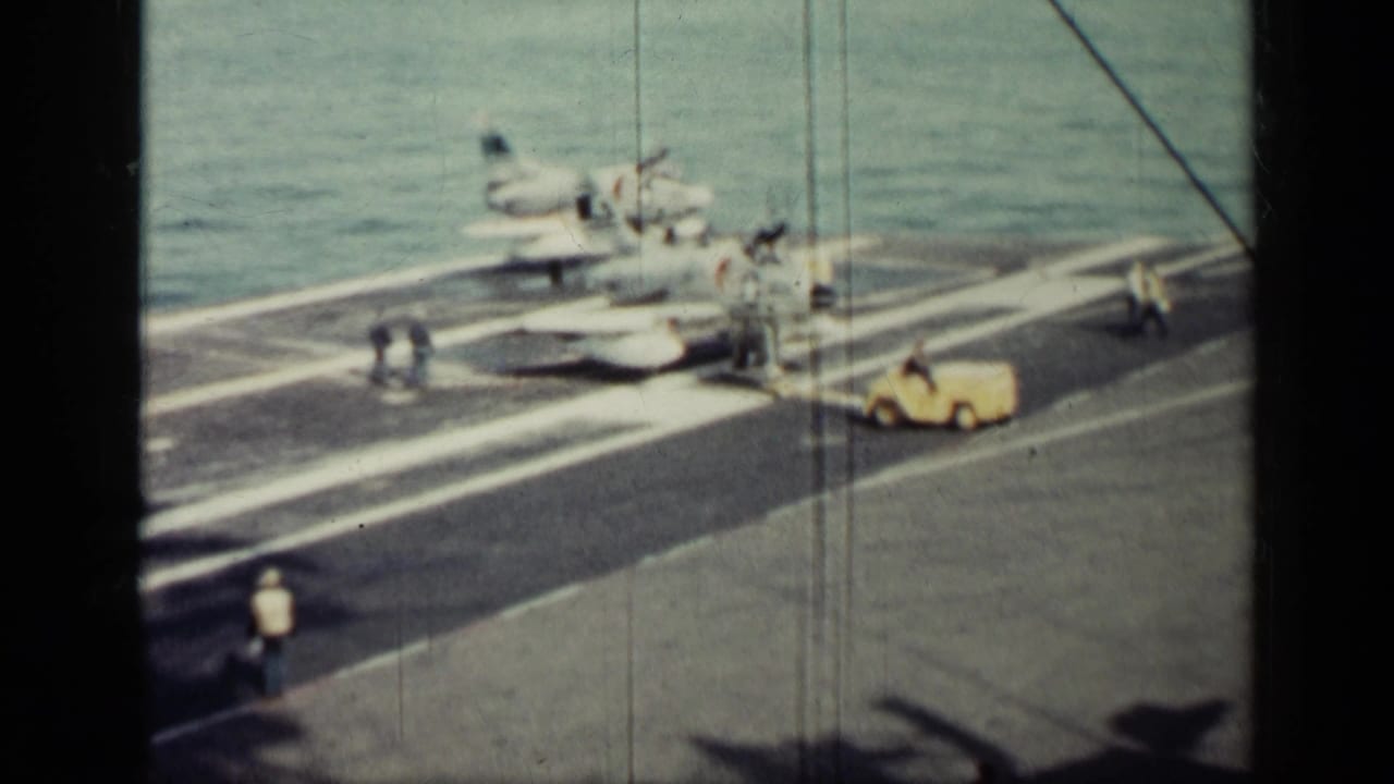 Propeller aircraft on an aircraft carrier deck with crew and a yellow service vehicle near the ocean.