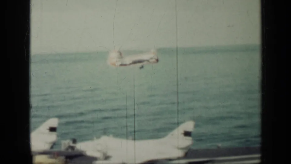 An aircraft is seen over the ocean beyond the carrier deck, with another aircraft partly visible in the foreground of this 8mm frame.
