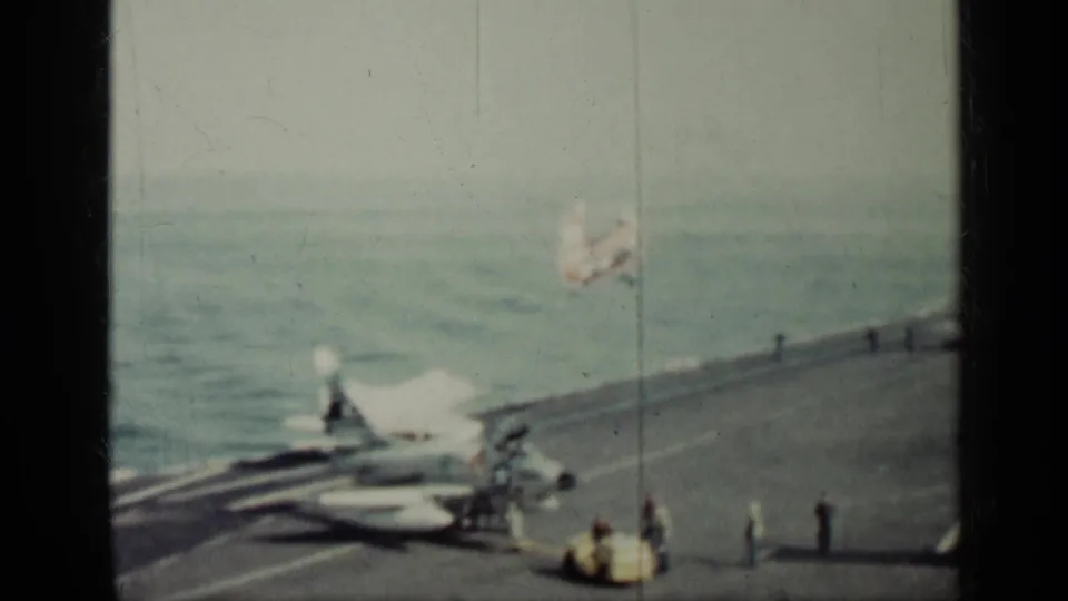 1962 Cuba 8mm frame showing carrier deck activity, a parked aircraft, distant deck figures, and a plane in flight over the sea.