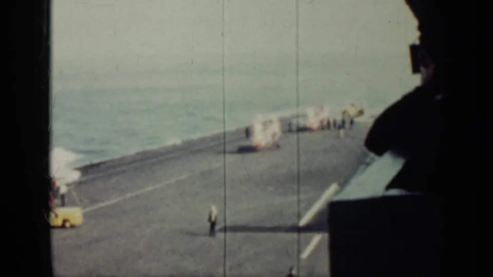 1962 Cuba-labeled frame showing a warship flight deck, distant crew near the helicopter area, ocean horizon, and yellow deck vehicle.