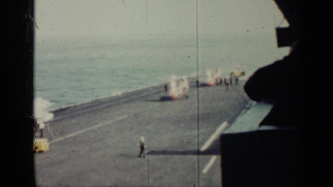 High view of a military ship flight deck with ocean, deck markings, small figures, and a dark foreground silhouette.