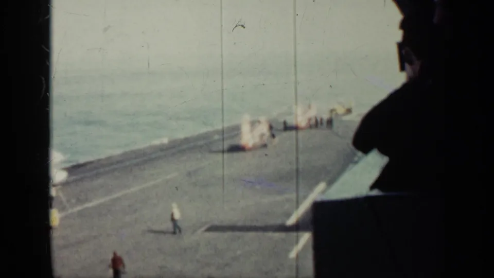 Cuba-linked 1962 home-movie frame showing a military ship flight deck, a walking figure, ocean, and deck gear.