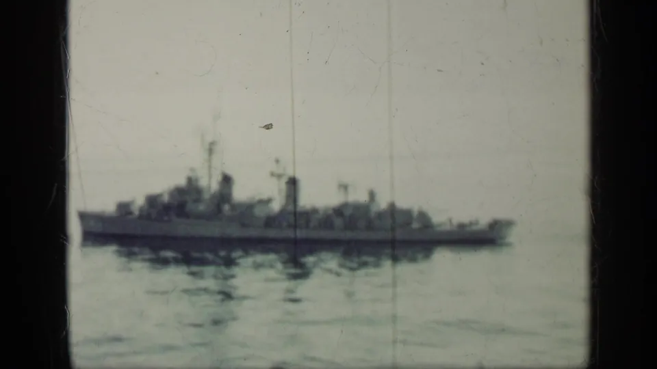 A gray naval vessel sits low on the horizon in a 1962 Cuba-linked home-movie frame with visible 8mm wear.