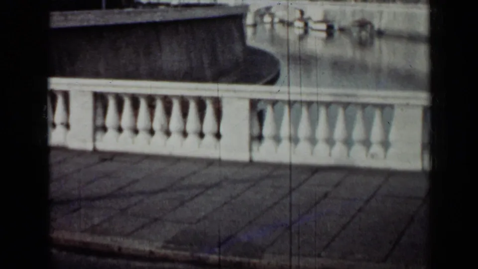 1962 Cuba 8mm frame of a white balustrade and paved promenade overlooking calm water and small moored boats.