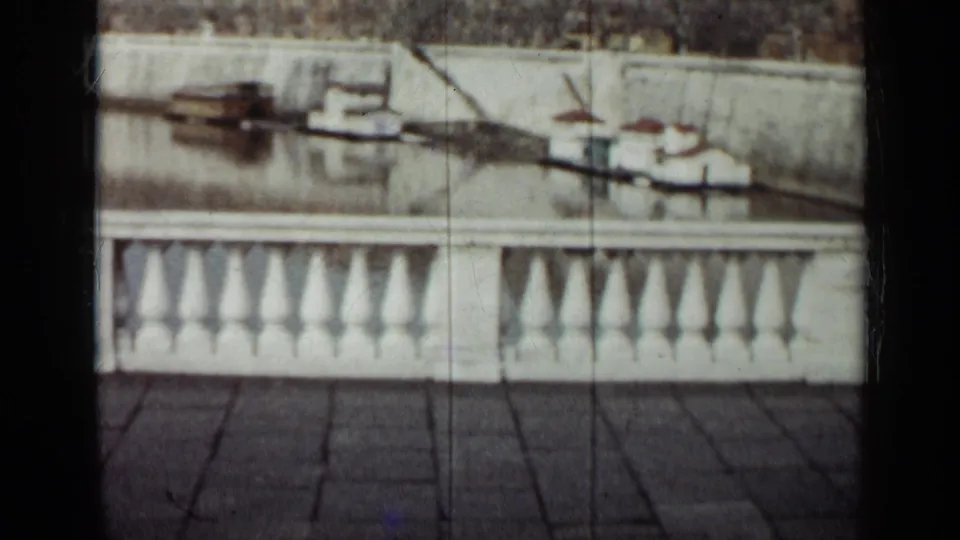 1962 Cuba 8mm frame showing a white balustrade in front of calm water and several small moored boats near a wall.