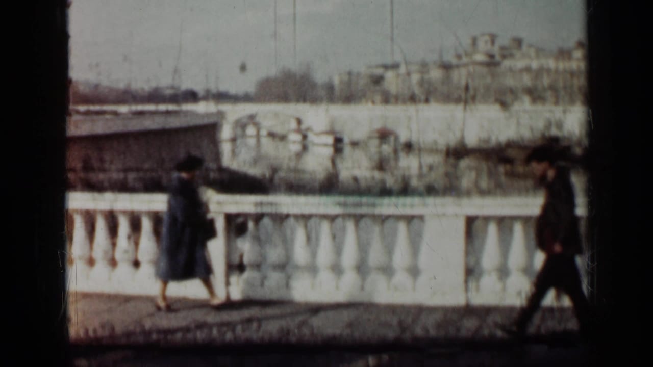 Pedestrians walk beside a white balustrade overlooking water or a lower roadway.