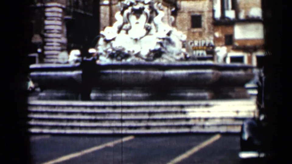 1962 Cuba street view with an ornate stone fountain, shuttered buildings, road markings, and partial shop lettering in the background.