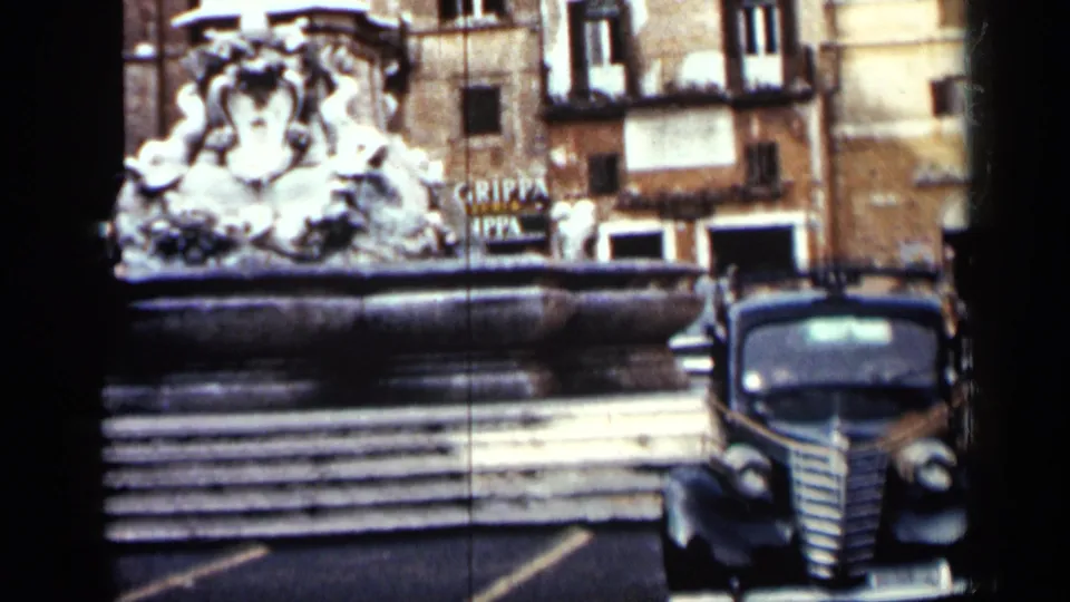 1962 Cuba home-movie frame showing an older car beside a stone fountain, with apartment facades and partial signage behind.