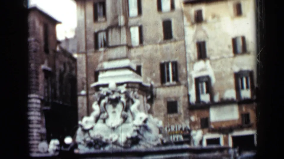 1962 Cuba urban view with a sculpted fountain, tall masonry buildings, shuttered windows, and partial commercial lettering.