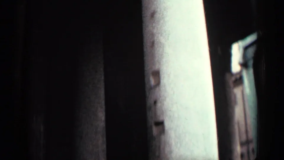 Handheld 8mm frame of a shadowed architectural passage with a pale vertical column or wall edge at right.