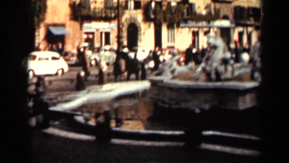 1962 Cuba public square view with a stone fountain in the foreground, pedestrians and cars beyond, and warm facades with balconies.