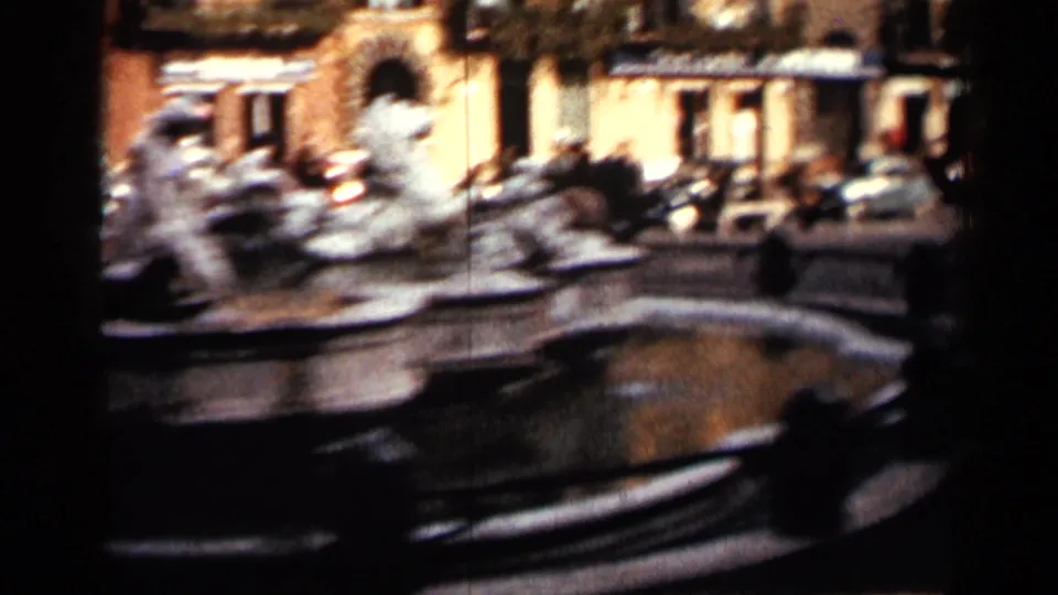 Handheld 8mm view of a public fountain in Cuba, with blurred stone sculpture, reflective water, cars, pedestrians, and surrounding facades.