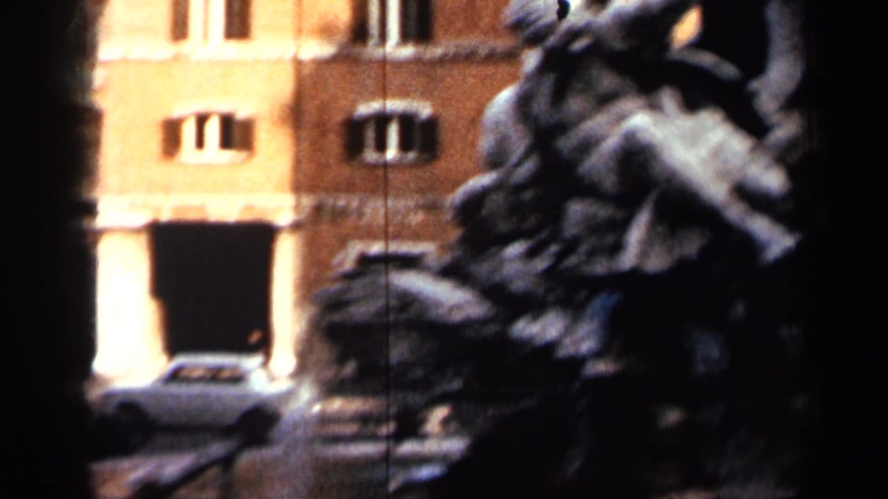 Dark sculptural fountain in front of sunlit yellow buildings with a white car behind it.