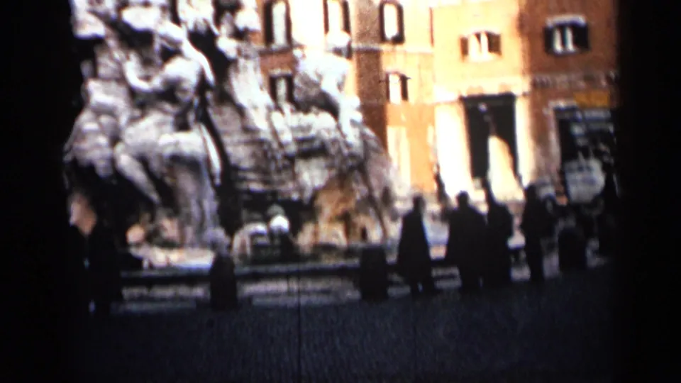 1962 Cuba home-movie view of a large public fountain beside buildings, with pedestrians crossing or pausing in the square.