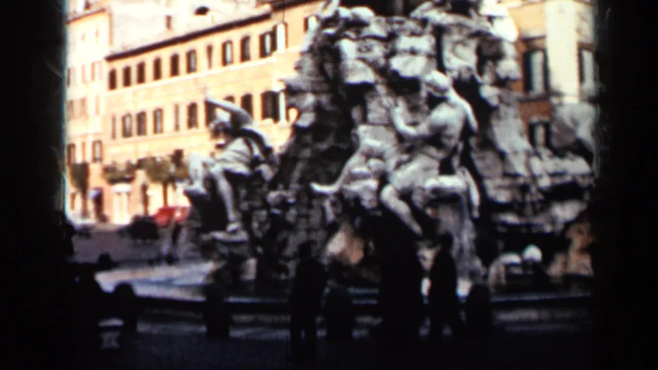 1962 Cuba frame showing the fountain from the side, with people near the stone base and yellow city buildings beyond.
