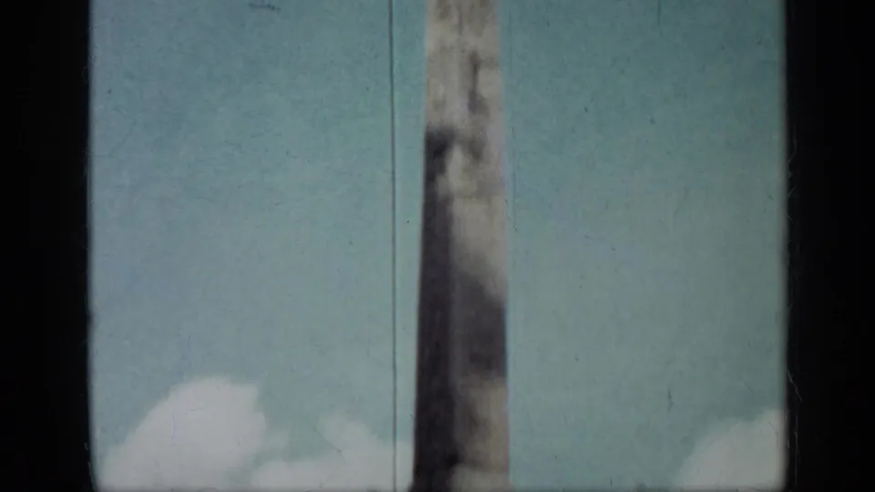A partial 8mm frame isolates the fountain obelisk against a pale blue sky with visible gate edge and film wear.