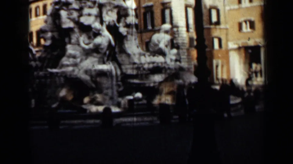 Handheld 8mm view of the fountain and nearby civilians, with city facades and dark frame edges around the scene.