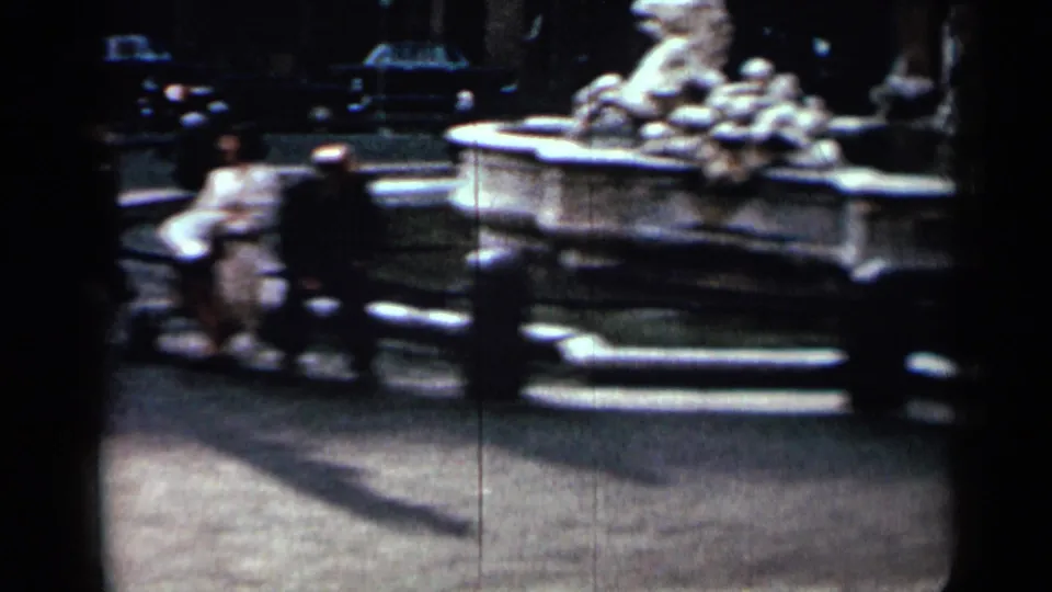 Soft 8mm shot of the fountain basin with sculpture, surrounding street activity, and civilians near the landmark.