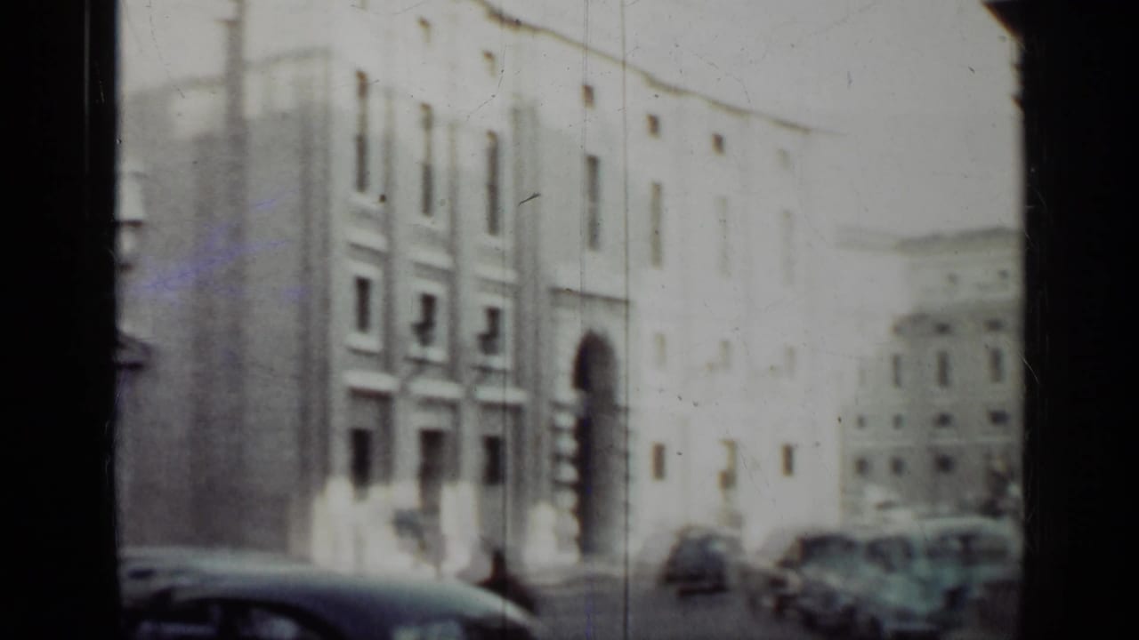 Soft view of a city street with pale buildings, parked cars, and a dark film edge.