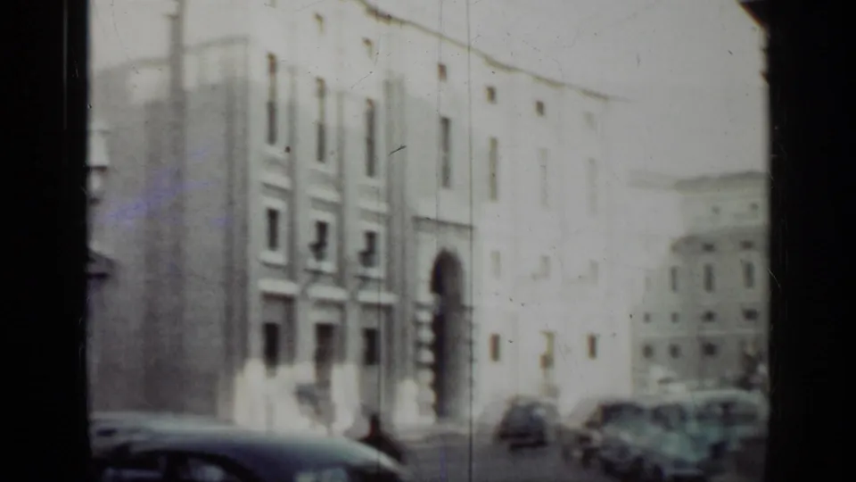 1962 Cuba home-movie frame showing pale urban buildings, a street with cars, and soft handheld 8mm exposure.