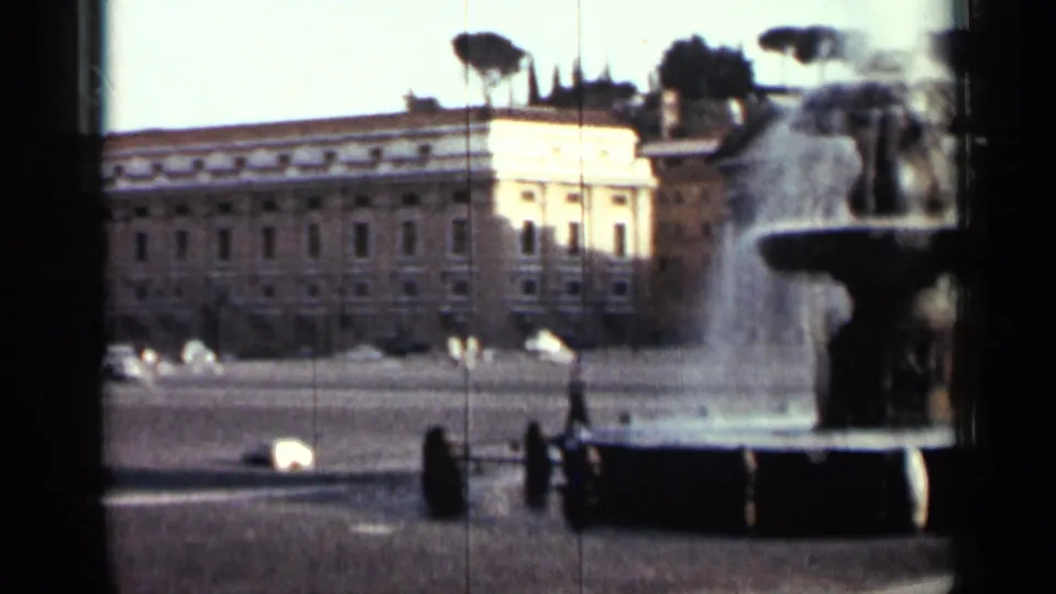 1962 Cuba home-movie frame showing a public fountain beside a broad square, formal buildings, cars, and distant people.