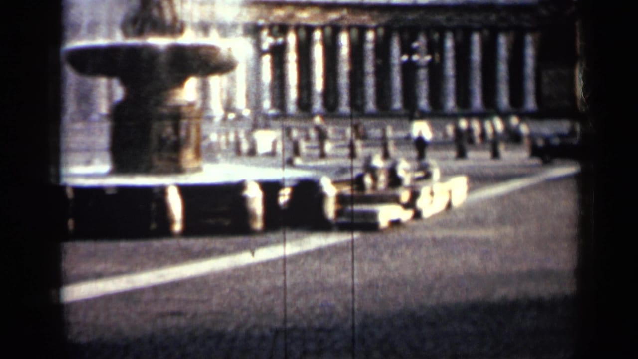 Large fountain in a paved square with a columned building and small pedestrians in the distance.