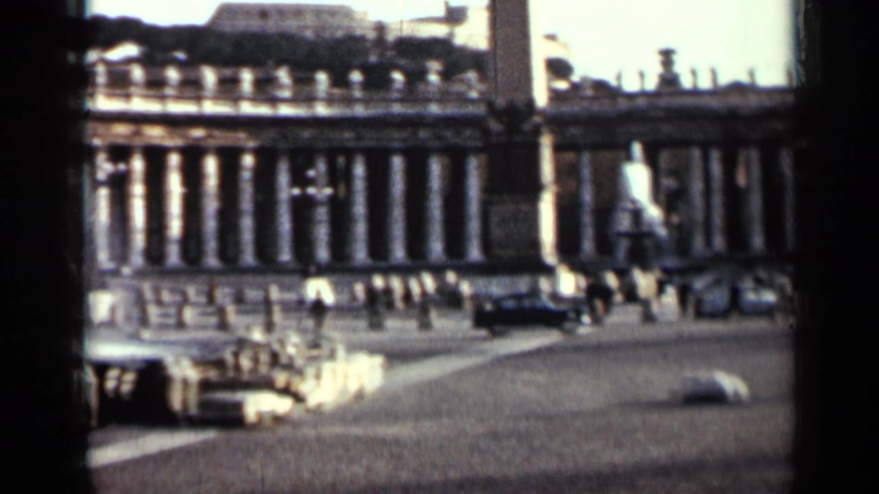 Wide colonnaded building across a cobblestone street with cars and dark foreground framing.