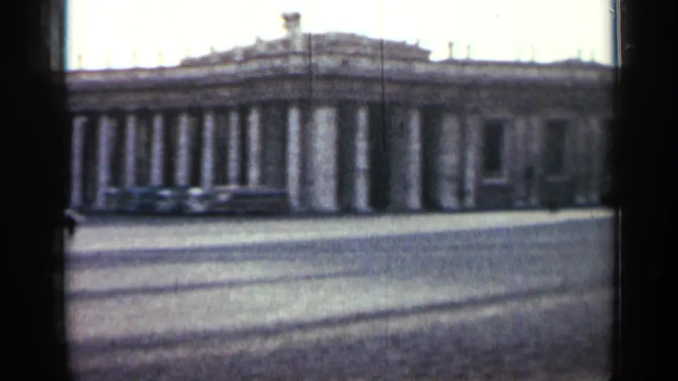 Soft 8mm frame of a Cuban landmark in 1962, with a long columned facade, open cobblestone street, and dark window-like framing.